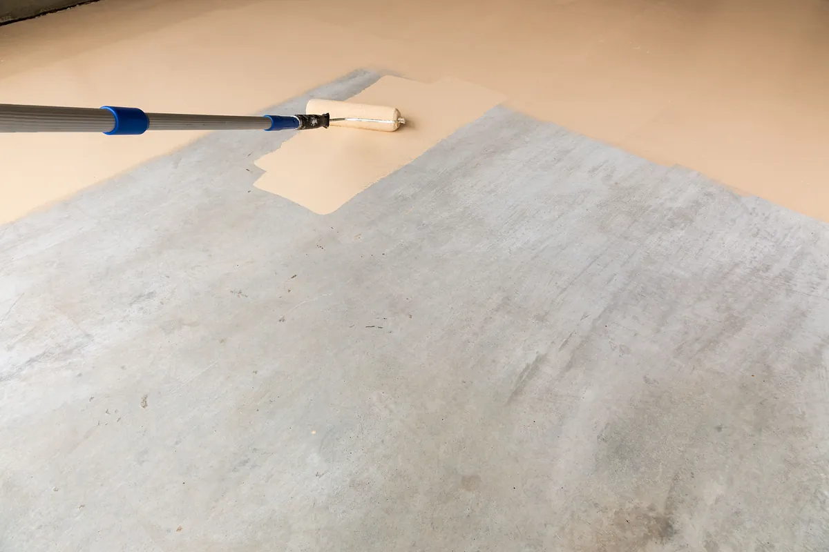 Inomhusmålning av golv. Bilden föreställer någon som målar ett grått betonggolv i en ljus beige färg.